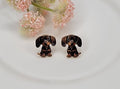 Small baby dachshund stud earrings with realistic black and brown enamel detailing, displayed on a white plate with soft pink flowers in the background.