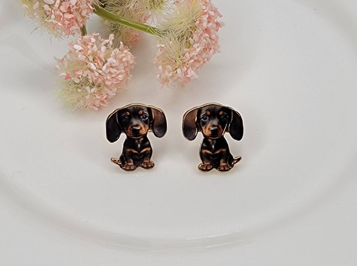 Small baby dachshund stud earrings with realistic black and brown enamel detailing, displayed on a white plate with soft pink flowers in the background.
