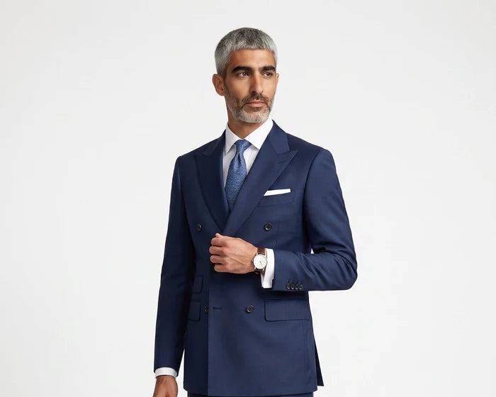 A man modeling a navy blue double-breasted suit with a white shirt, blue tie, and white pocket square, standing against a light gray studio background.