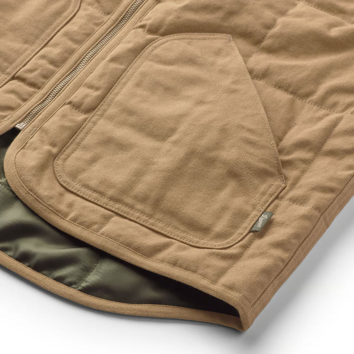 Close-up of a tan quilted jacket with a large front patch pocket, zipper closure, and a small metal brand tag, shown on a white background.