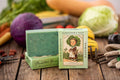 Two bars of Gardener's Soap with packaging on a wooden surface with vegetables in the background.