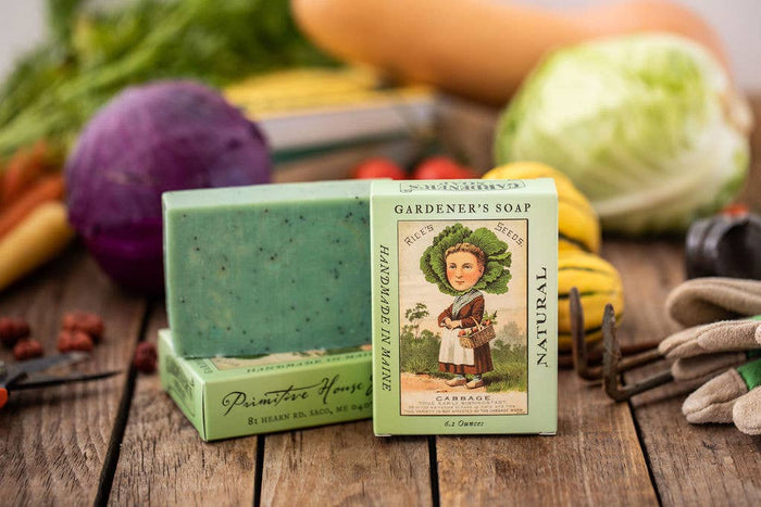 Two bars of Gardener's Soap with packaging on a wooden surface with vegetables in the background.