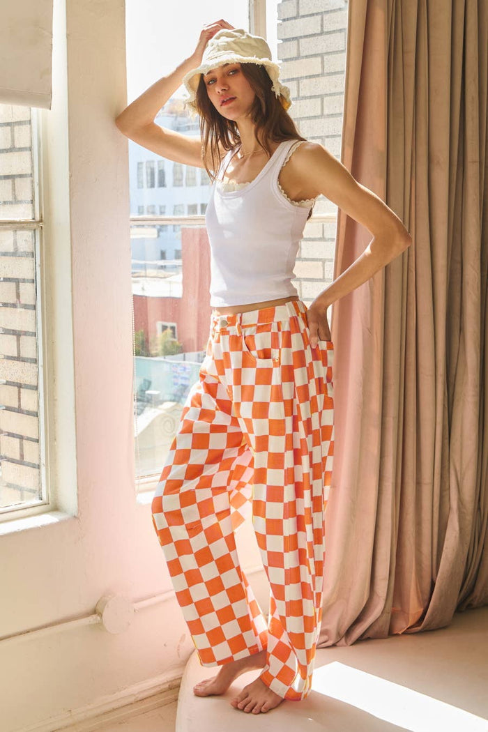 Woman wearing orange checkered barrel-leg denim pants styled with a white tank top, showcasing bold pattern and relaxed fit from The Pink Doxie Boutique.
