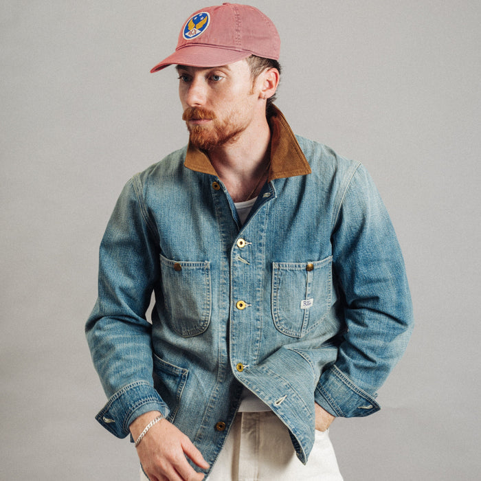 A man wearing a blue denim work jacket with a brown corduroy collar, paired with off-white trousers and a dusty pink baseball cap, photographed against a neutral gray studio background.