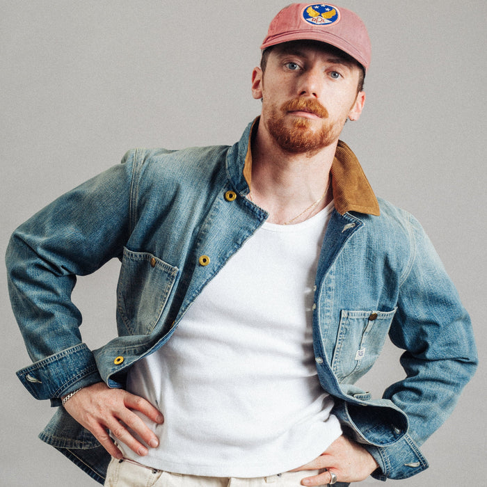 A man wearing a dusty pink baseball cap, a blue denim jacket with a brown corduroy collar over a white t-shirt, and cream-colored pants, posing against a solid gray studio background.