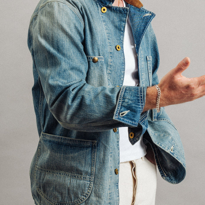 A person wearing a blue denim jacket with gold-tone buttons over a white shirt and light beige pants, photographed against a plain gray studio background.