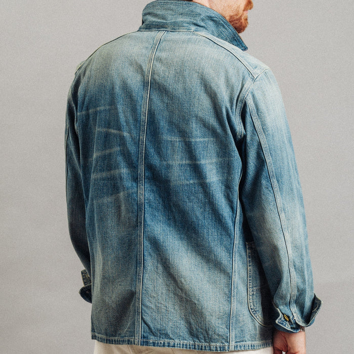 A man wearing a light blue washed denim jacket, shown from the back, paired with off-white trousers against a neutral gray studio background.