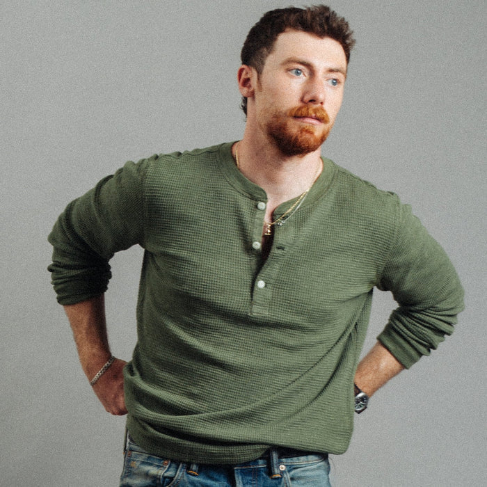 A man with a red beard wearing an olive green ribbed long-sleeve henley shirt and blue jeans, posing with hands on hips against a solid gray background.