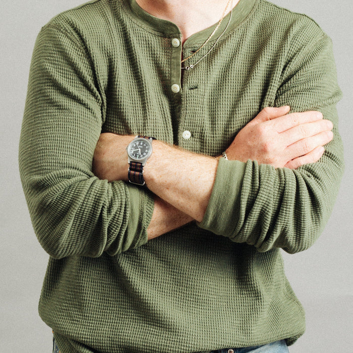 A man wearing an olive green waffle-knit long-sleeve henley shirt and blue jeans, with his arms crossed. He is wearing a brown leather strap watch on his left wrist.