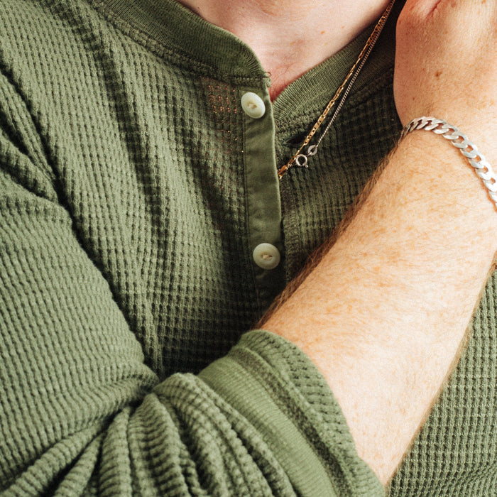 Close-up of a person wearing an olive green waffle-knit long-sleeve henley shirt, with the collar open, buttons visible, and one arm raised wearing a silver chain bracelet.