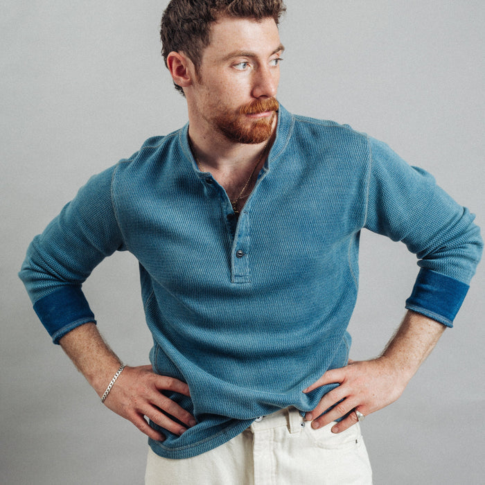 A man with a reddish beard wearing a textured blue long-sleeve henley shirt and off-white pants, posing with hands on hips against a solid gray studio background.