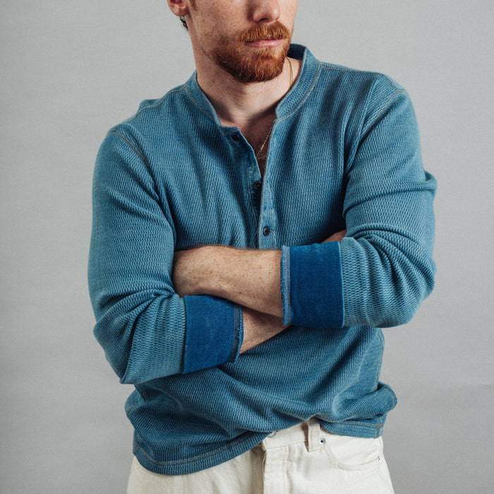 A man with red hair and a beard wearing a textured blue henley shirt with contrasting dark blue cuffs, posed with arms crossed against a solid gray background.