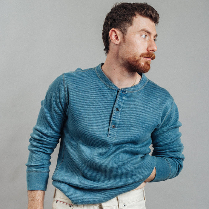 A man wearing a teal blue ribbed long-sleeve henley shirt and off-white jeans, standing against a plain gray studio background.