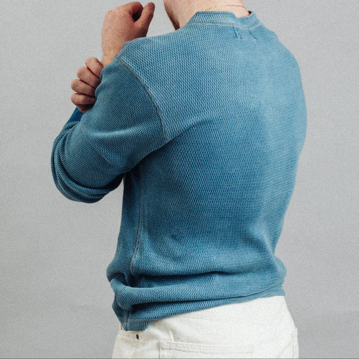 A man wearing a textured blue knit crewneck sweater, shown from the back and side against a plain gray studio background.