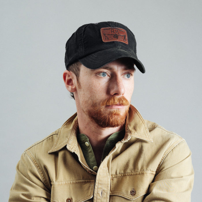A man with a red beard wearing a black baseball cap with a brown patch and a tan button-up shirt, photographed against a plain light gray background.