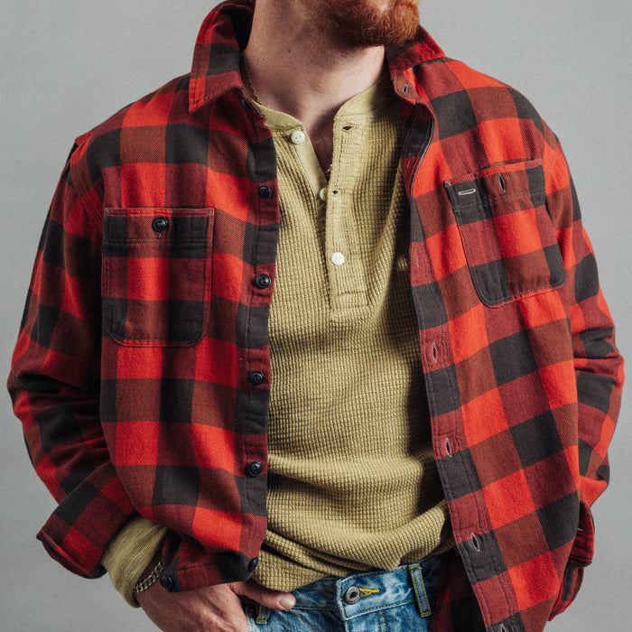 A man wearing a red and black buffalo plaid flannel shirt over a muted olive green henley shirt, paired with blue jeans, against a neutral gray studio background.
