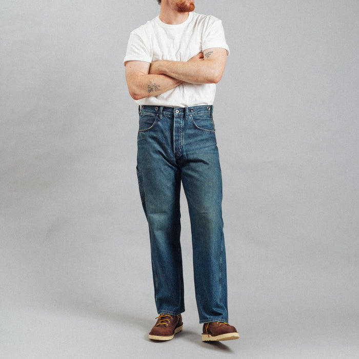 A man wearing a white t-shirt, blue jeans, brown shoes, and a black baseball cap stands with arms crossed against a plain gray studio background.