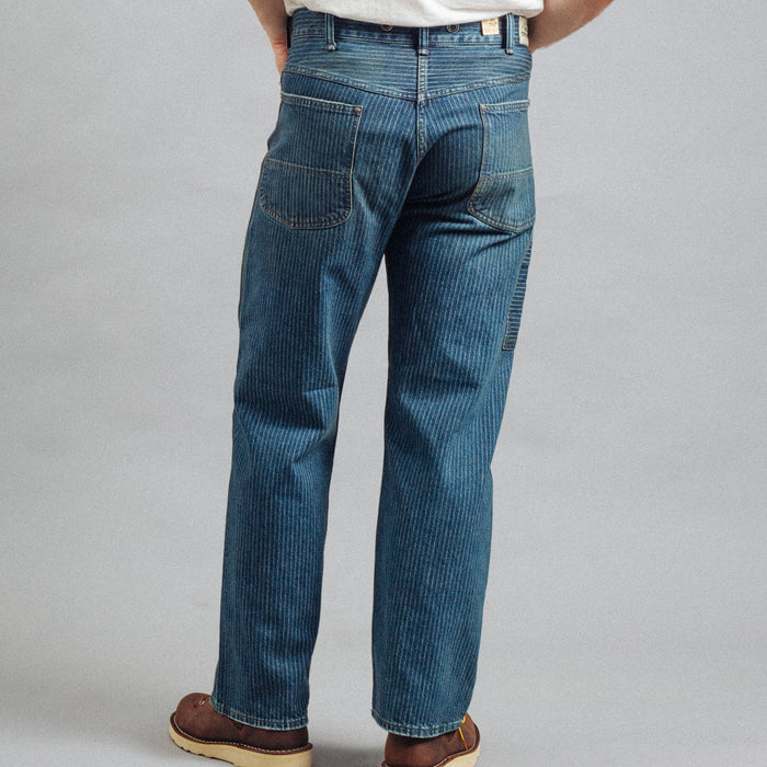 A person shown from the back wearing blue denim jeans, a white t-shirt, and brown shoes against a plain gray studio background.
