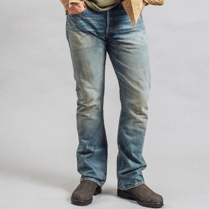A person wearing light-wash blue denim jeans with a tan overshirt and dark brown boots, shown from the waist down against a plain light gray studio background.
