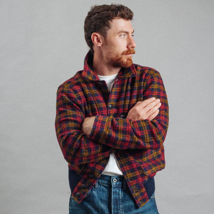 A man wearing a red and yellow plaid flannel shirt over a white t-shirt and blue jeans, standing with arms crossed against a plain gray background.