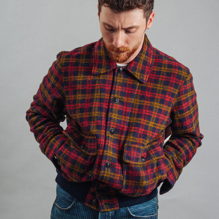 A man wearing a red, yellow, and blue plaid button-up overshirt jacket over a dark shirt and blue jeans, photographed against a plain gray studio background.