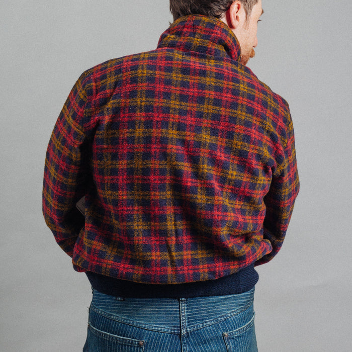 A man viewed from behind wearing a red, yellow, and dark blue plaid fleece jacket and blue jeans against a plain gray studio background.